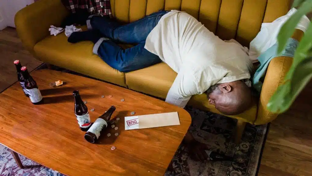 A man lying face down on a couch with beer bottles and scattered coins on a coffee table.