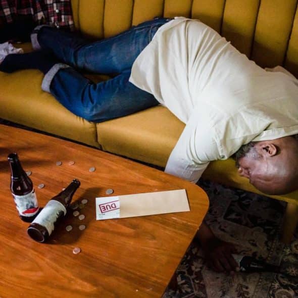 A man lying face down on a couch with beer bottles and scattered coins on a coffee table.