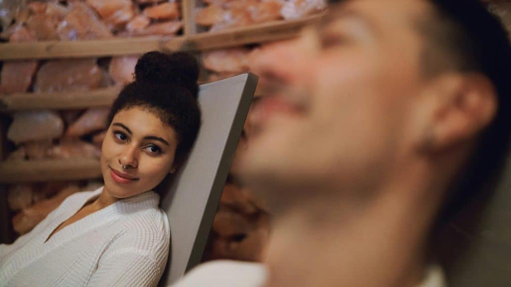 A woman in a white robe smiling while looking at a man relaxing nearby.