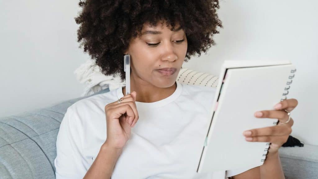 A woman sitting on a couch holding a notebook and a pen in a thoughtful pose.