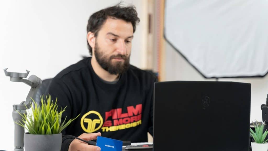 Bearded man working at a laptop, holding a small blue **Samsung** portable SSD.