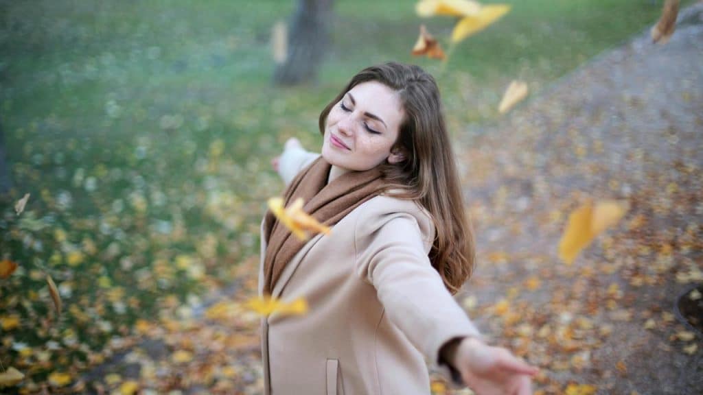 A woman in a beige coat smiling with arms outstretched as autumn leaves fall around her.