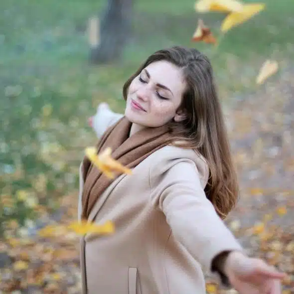 A woman in a beige coat smiling with arms outstretched as autumn leaves fall around her.
