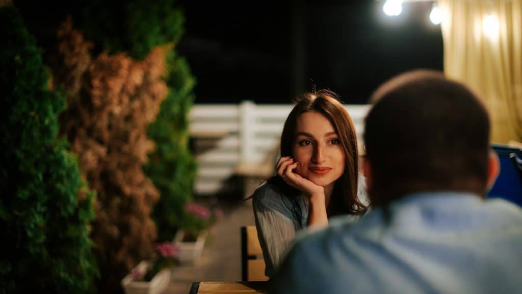 A smiling young woman rests her chin on her hand while looking at a man in the foreground, sitting at a table in a dimly lit outdoor patio with lights and trees.