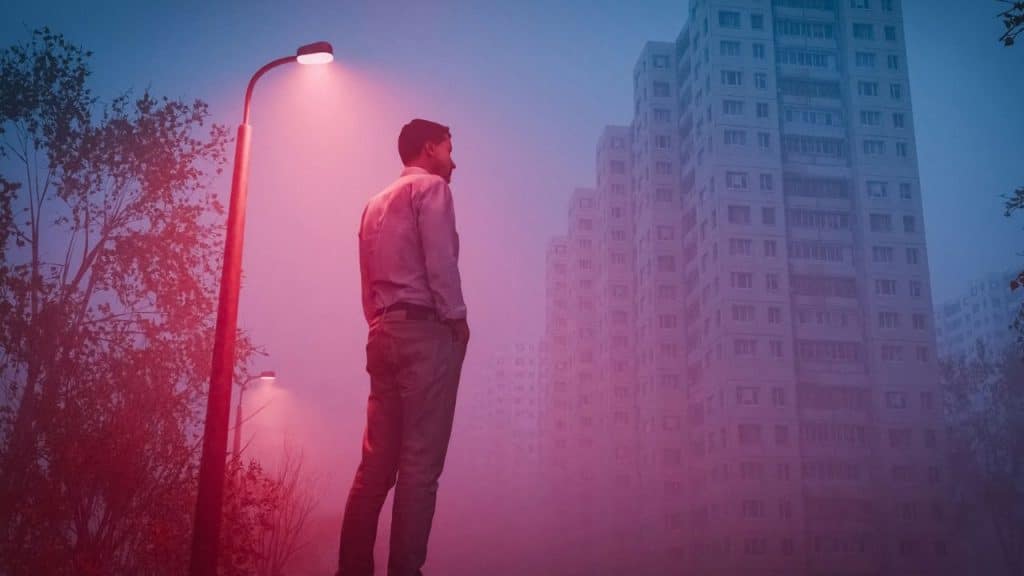 A man stands alone at night under a red-lit streetlamp next to tall, fog-obscured buildings.