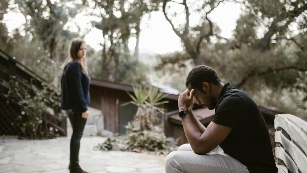 A man sitting with his head in his hands while a woman stands nearby looking upset.