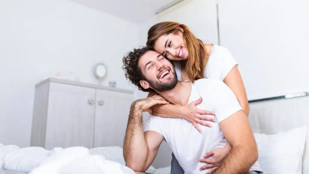 A man and a woman in white shirts are smiling and embracing in a bright room.