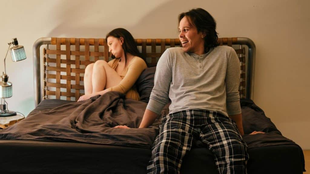 A smiling man sits on a bed while a disgruntled woman sits turned away from him in the background.