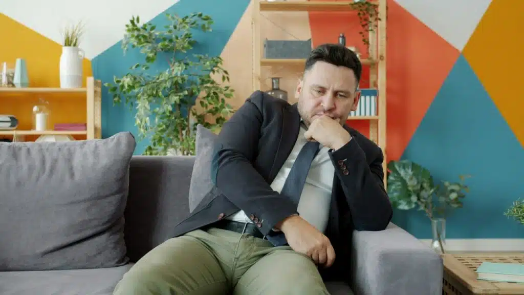 A man in a suit jacket is sitting thoughtfully on a couch in a colorful room.