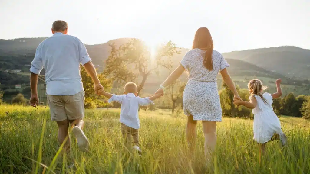 A family of four holding hands, walking away into a sunny field at sunset.