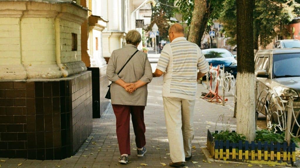 Elderly couple viewed from behind walking arm-in-arm down a sidewalk in a city.