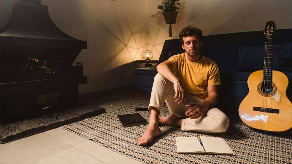 A man is sitting on the floor by a fireplace and a guitar, looking at the camera.