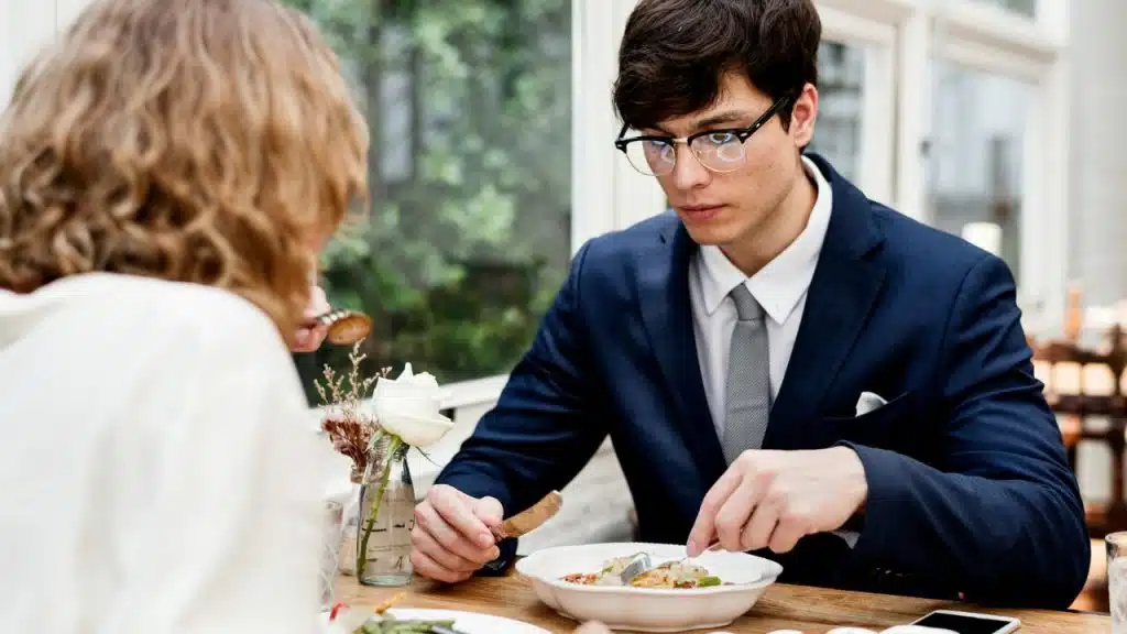 A serious man in a blue suit and glasses eats at a table with a blurred woman opposite him.