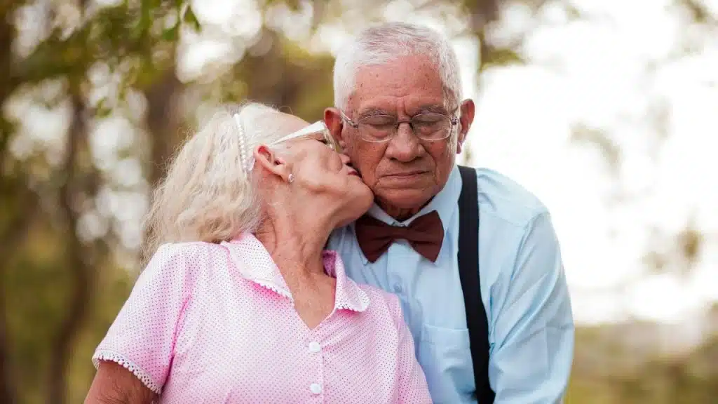 An elderly woman in pink kisses an elderly man in a bow tie and suspenders on the cheek.