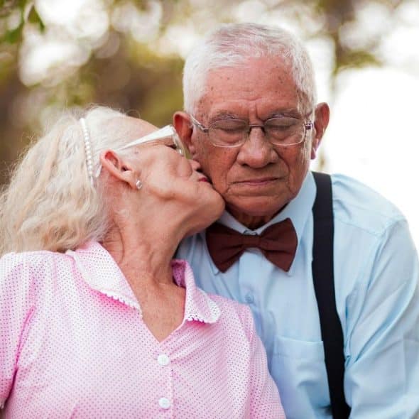An elderly woman in pink kisses an elderly man in a bow tie and suspenders on the cheek.