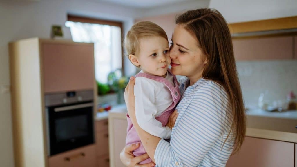 A mother in a striped shirt holds a young child in a pink overall in a kitchen.