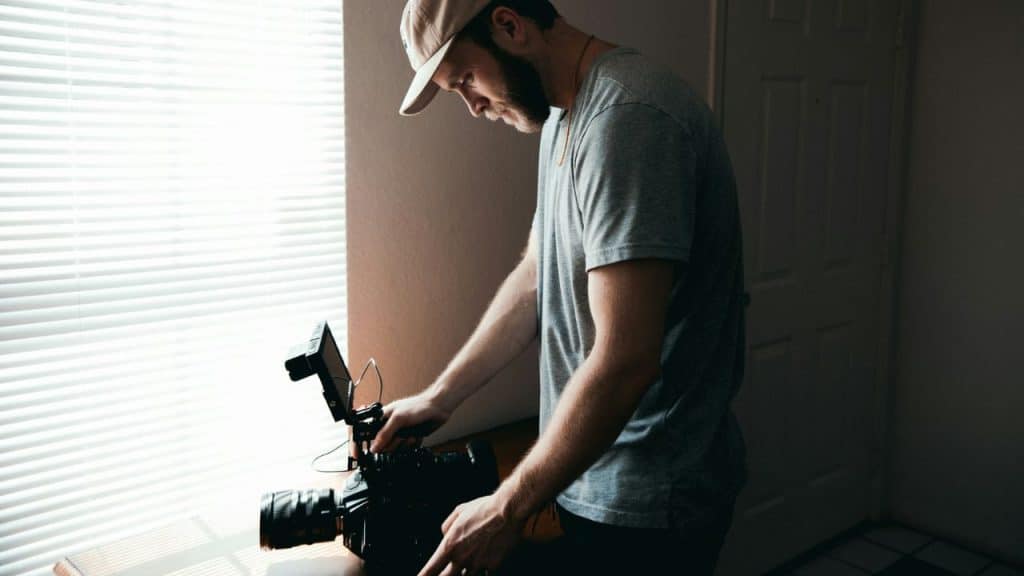 A bearded filmmaker in a cap is adjusting a professional camera by a window.