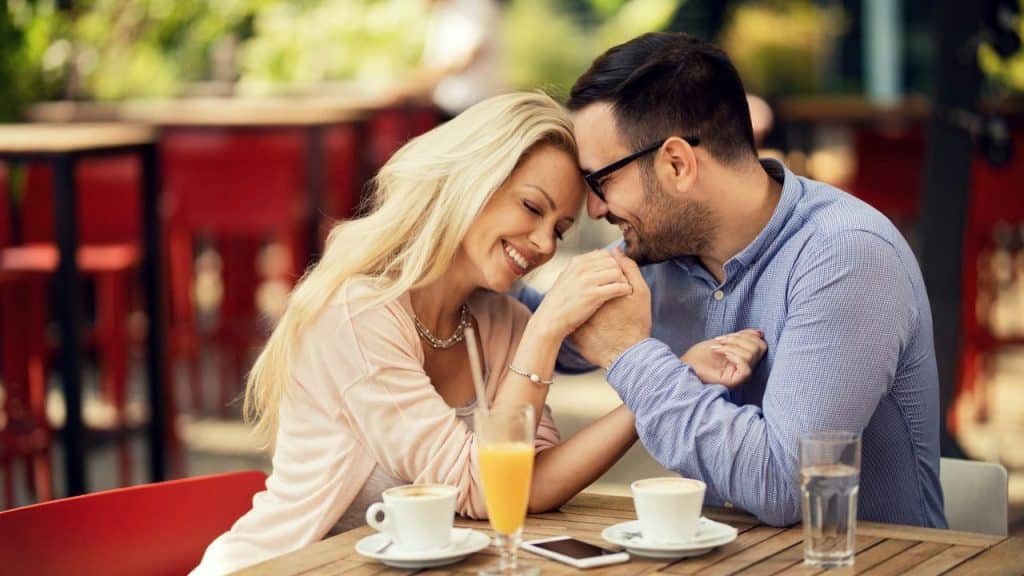 Smiling couple holding hands and leaning in close at an outdoor cafe table.