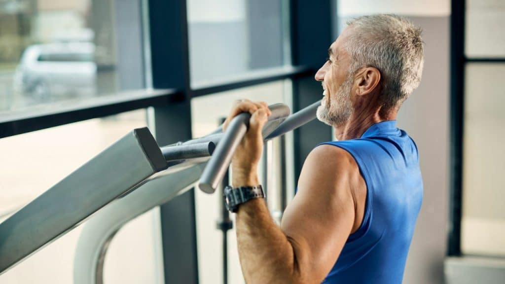 Smiling older man in a blue tank top doing a pull-up or other resistance exercise at the gym.
