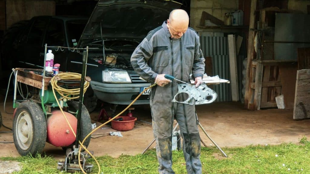 An older, bald mechanic in a greasy jumpsuit is working on a metal part.