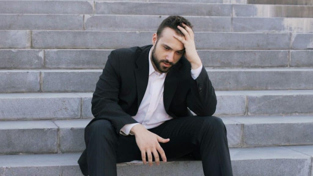 A man in a black suit sits on concrete steps with his hand pressed to his head.