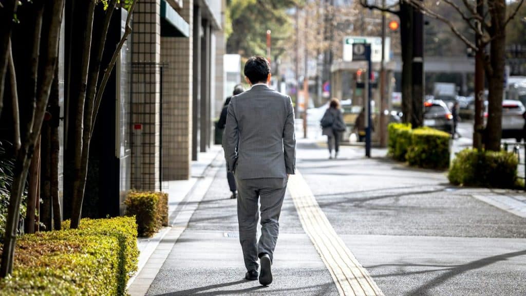 Man in a gray suit walks away down a city sidewalk lined with trees and bushes.