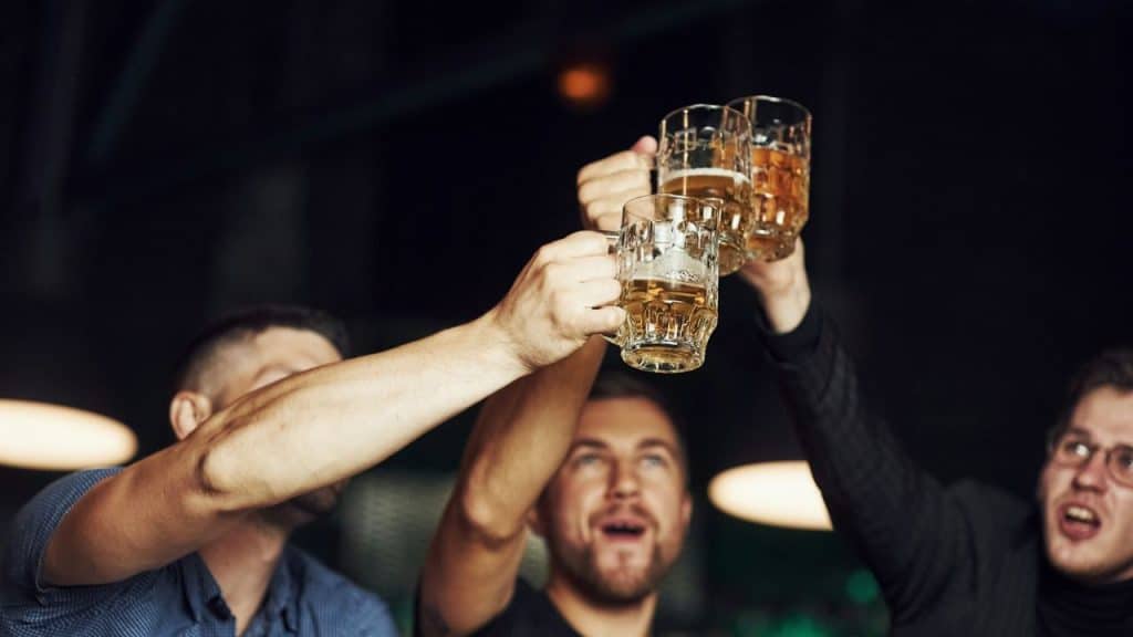 Three men raise glass mugs of beer in a dark bar, cheering and toasting together.