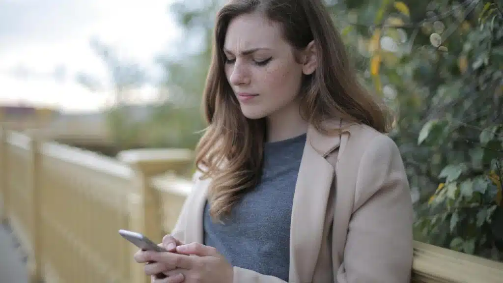 A woman standing outdoors looking at her phone with a concerned expression.