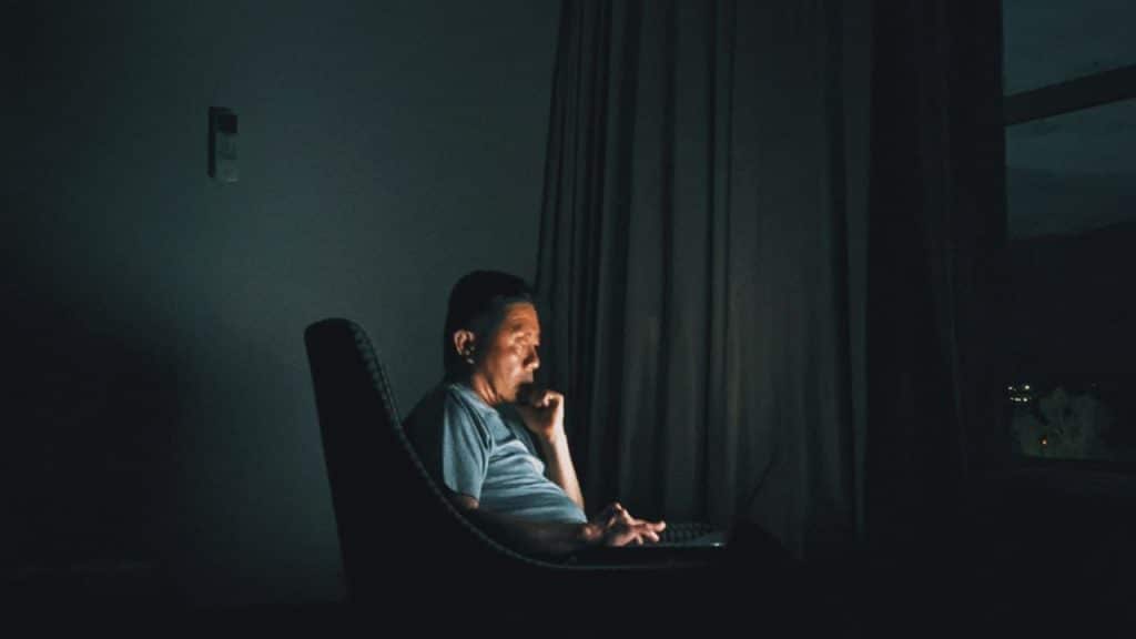 Man on a chair in a dark room illuminated by the glow of a laptop screen.