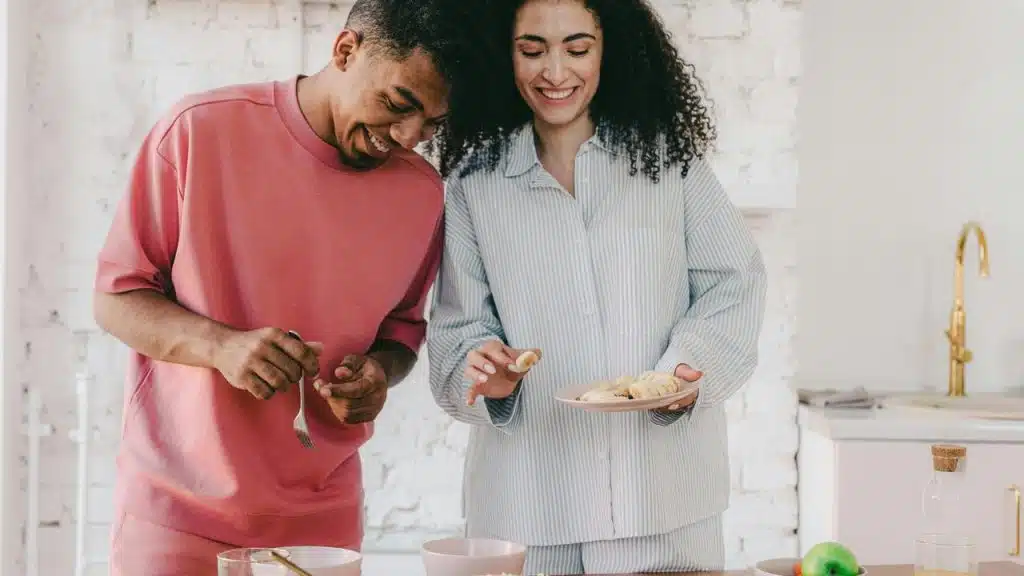A couple smiling while preparing food together in a kitchen.