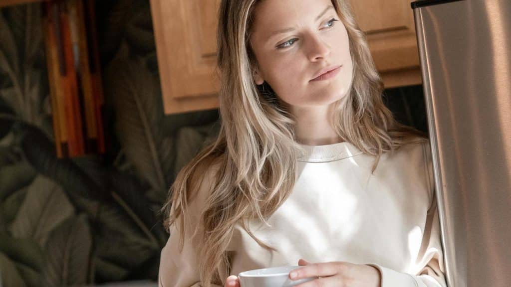 A woman standing in a kitchen holding a cup and looking thoughtful.