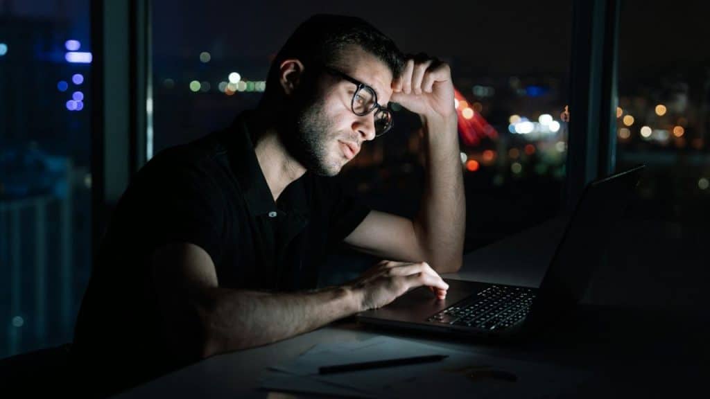 A man with glasses works on a laptop late at night in a dark room.