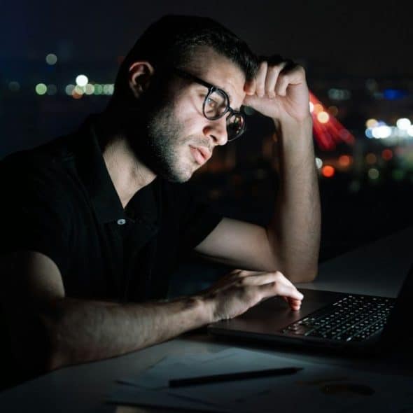 A man with glasses works on a laptop late at night in a dark room.