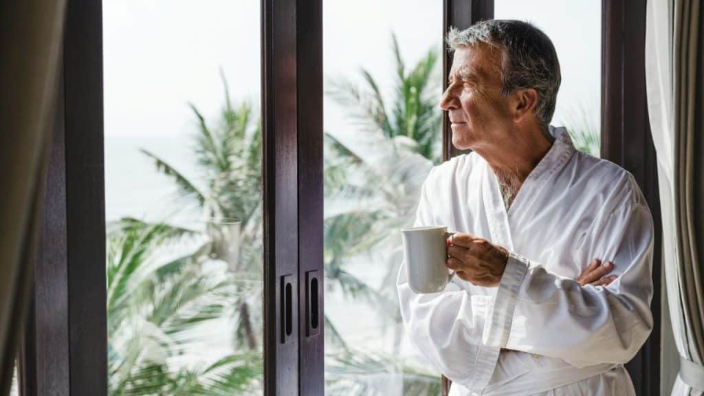 Older man in a white robe holding a mug, looking out a large window at palm trees and the ocean.