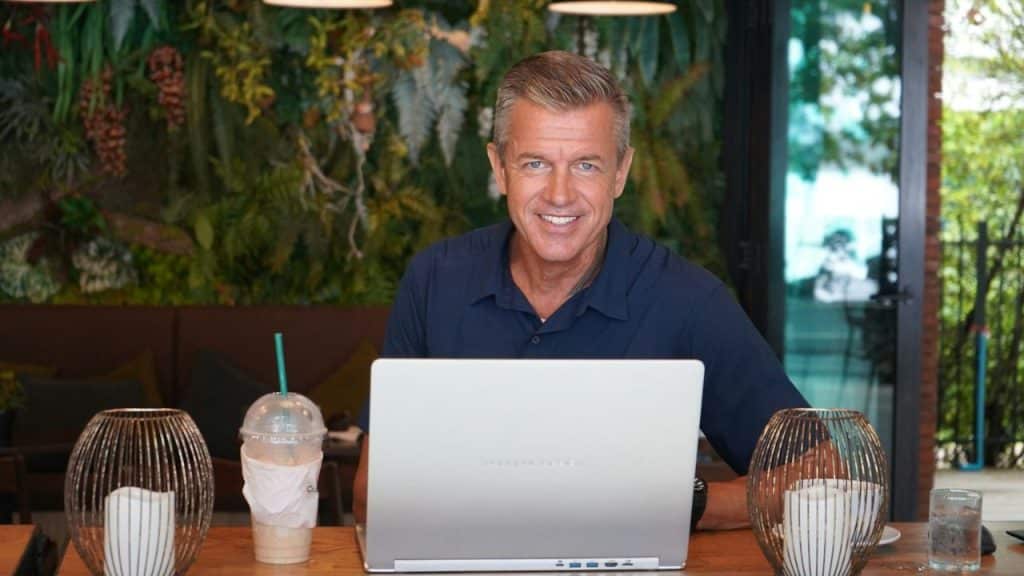 Smiling man sitting behind a laptop at a wooden table in a leafy cafe.