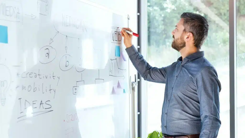 Male in a blue shirt with a beard writing on a whiteboard with a red marker.