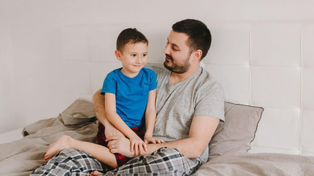 Man and young boy in pajamas sitting together on a bed, looking at each other.