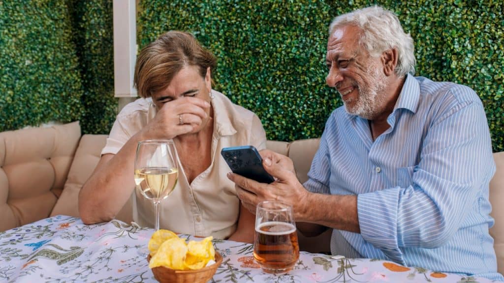 A couple sitting at a table with wine glasses with the man showing his wife something on his mobile while she laughs.