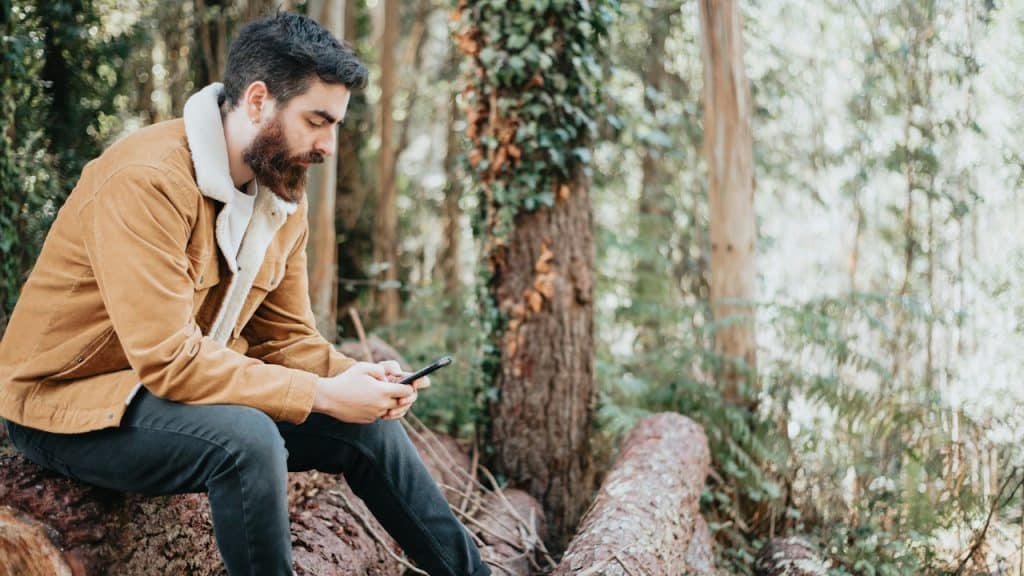A man sitting on a log in the woods while using his phone.