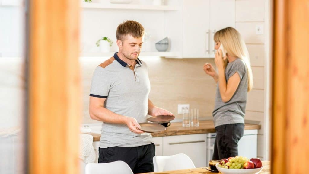A man is holding plates in a kitchen while a woman is talking on a cell phone in the background.