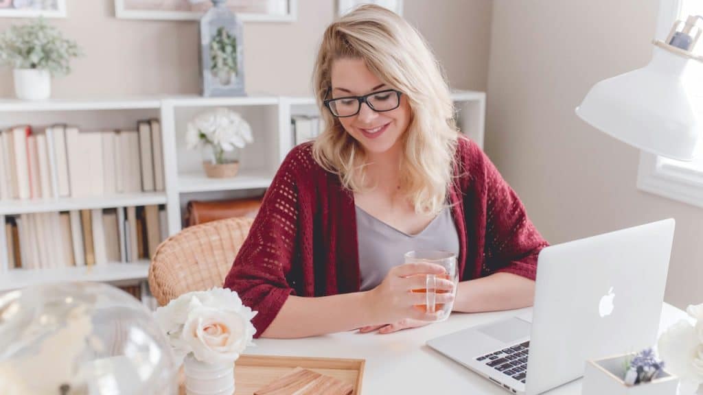 A smiling blonde woman wearing glasses working on a laptop at a clean home office desk.