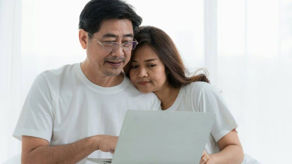 An older man wearing glasses and a younger woman are cuddling and looking at a laptop together.
