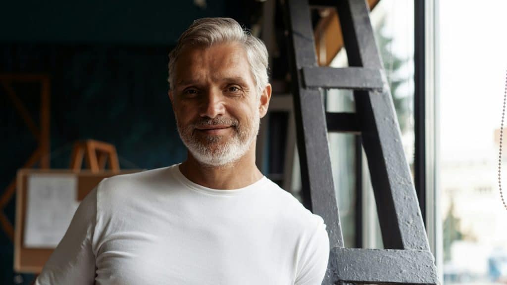 A mature man with gray hair and a white beard smiles directly at the camera while leaning against a dark metal structure indoors.