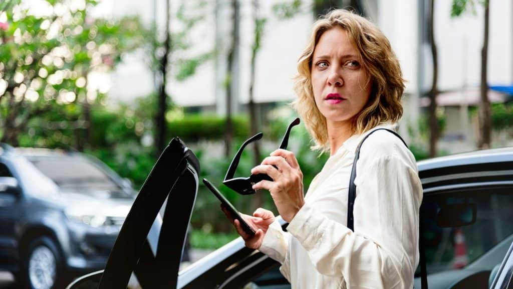 A woman with wavy blonde hair stands by an open car door, holding sunglasses and a phone, looking off to the side.