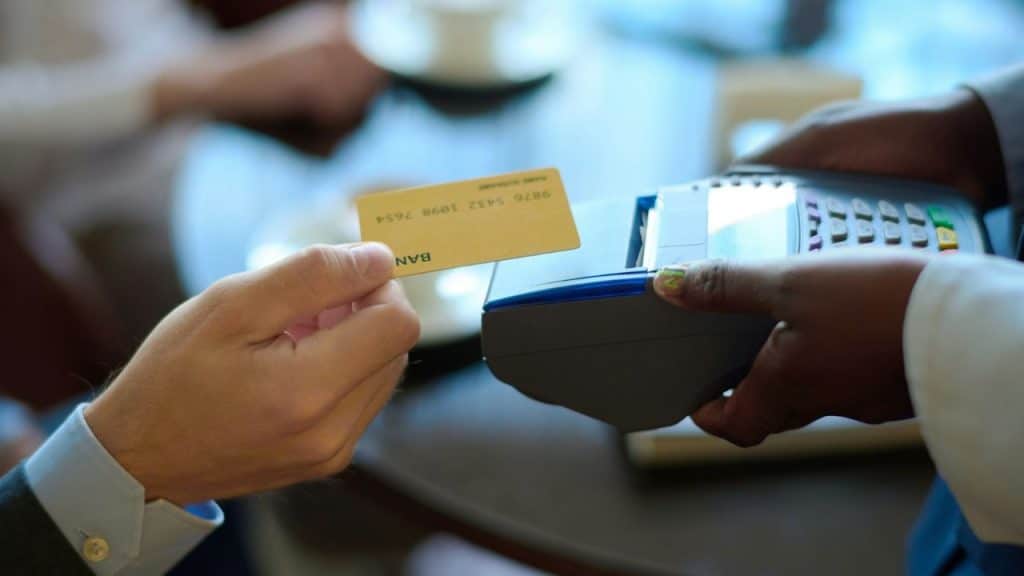 A man's hand is about to tap a gold credit card on a payment terminal held by another person.