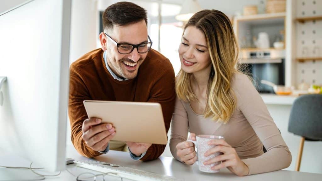 A smiling couple looks at a tablet while sitting at a white desk in a well-lit kitchen.