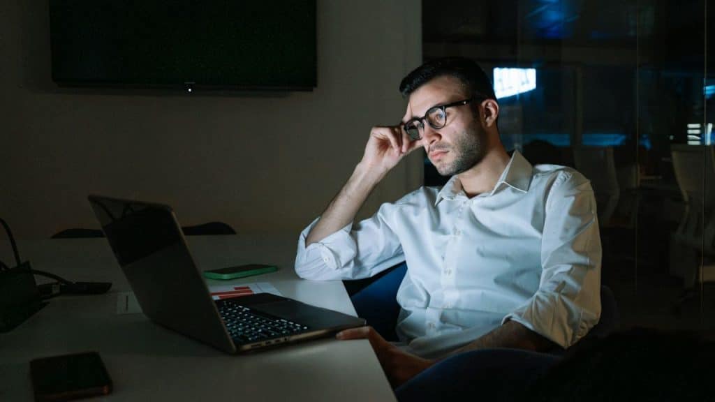 A serious man in a white shirt and glasses is intensely focused on his laptop in a dark room.