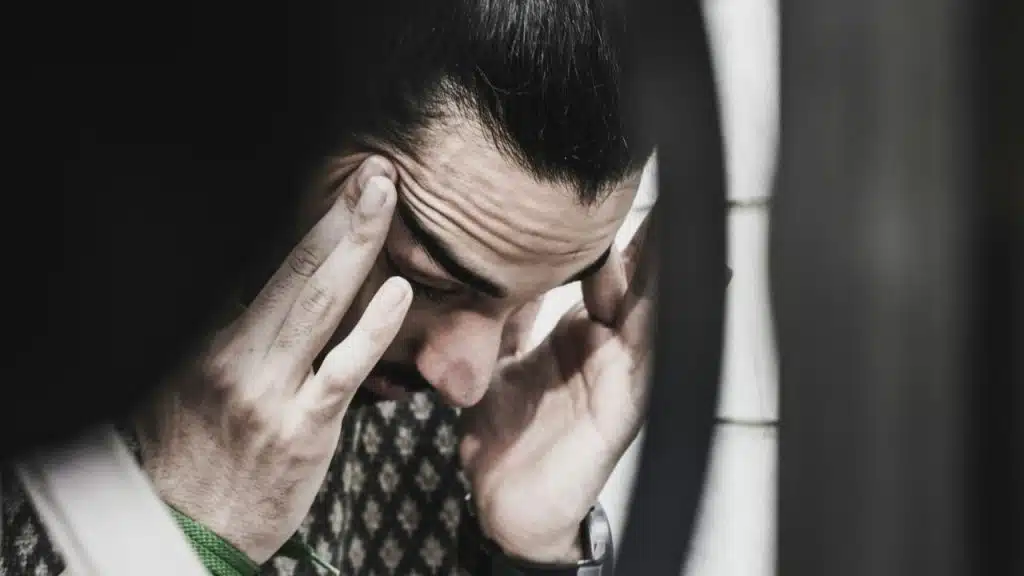 Close-up of a man with his eyes closed, pressing his hands to his temples.