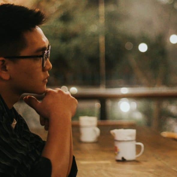 A man wearing glasses sits at a wooden table with his chin resting on his hand, looking away from the camera.