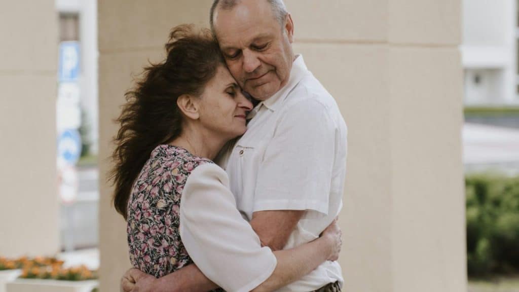 Older man in white shirt hugging an older woman in a floral dress.